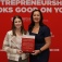 Amy Monin (left) and Hadar Borden hold an award while standing in front of a banner that contains sponsorship info at the conference. 