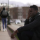 A young African-American man seated on a bench with his hand to his head as a group of people walk away in the background. 