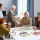 Three older adults sit at a table while one stands. An aide looks on. 