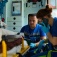 Two EMTs in an ambulance with a patient, one EMT uses a cell phone to communicate with the hospital. 