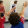 Photo shows the backs of two children raising their hands with a child and instructor in background in a classroom. 