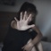 A woman, hugging her knees while sitting on a bed, holds her hand up to fend off another person. 