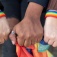 Close up of three people's fists; two are wearing rainbow bracelets and one is holding a rainbow flag. 